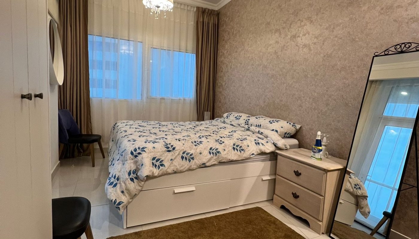 A neatly organized bedroom with a single bed, brown rug, large mirror, bedside table, and a window with beige curtains. A chair and wardrobe are also visible.