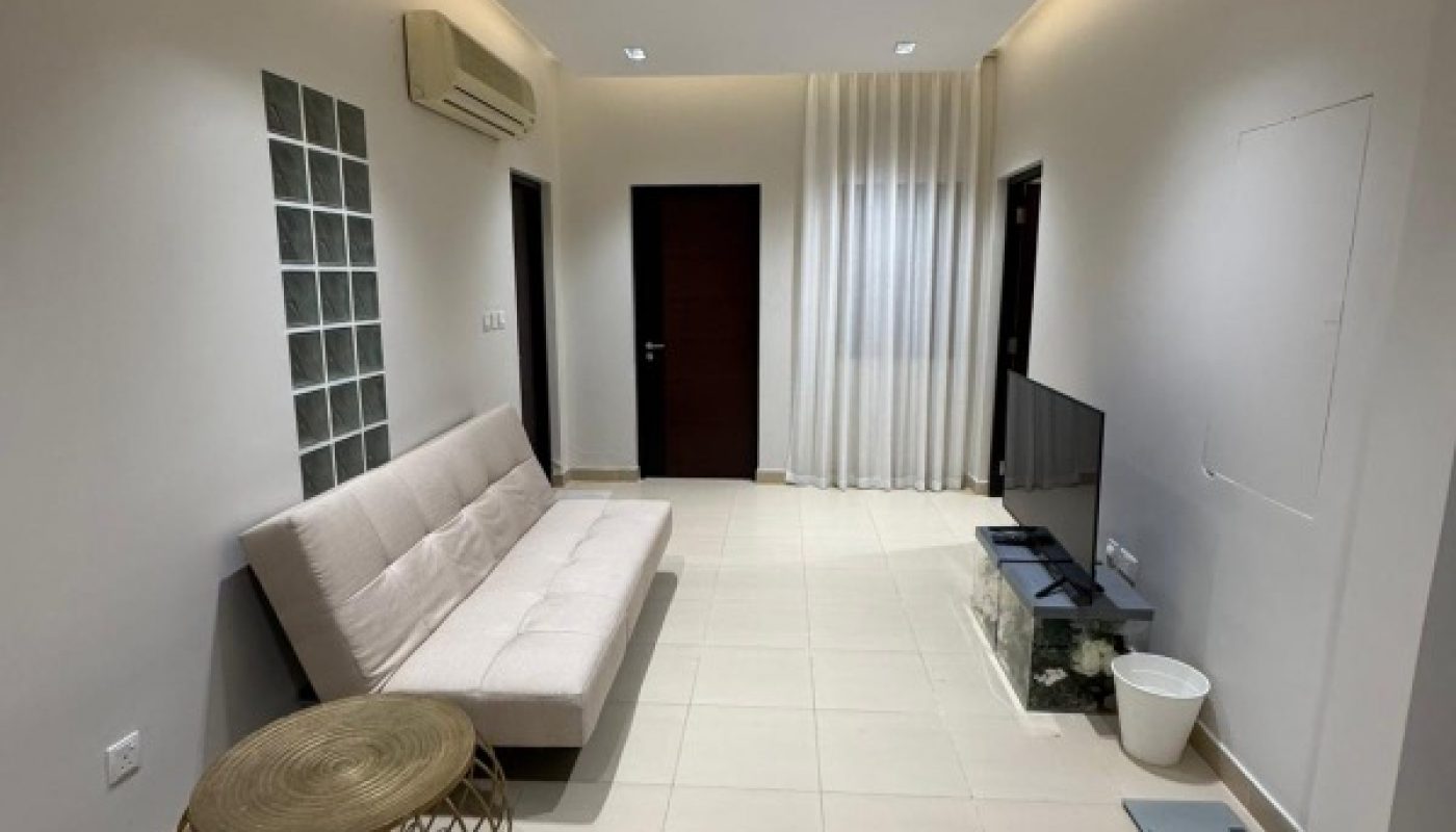 A minimalist living room with a beige sofa, round wicker table, TV on a glass stand, tile flooring, and white walls.