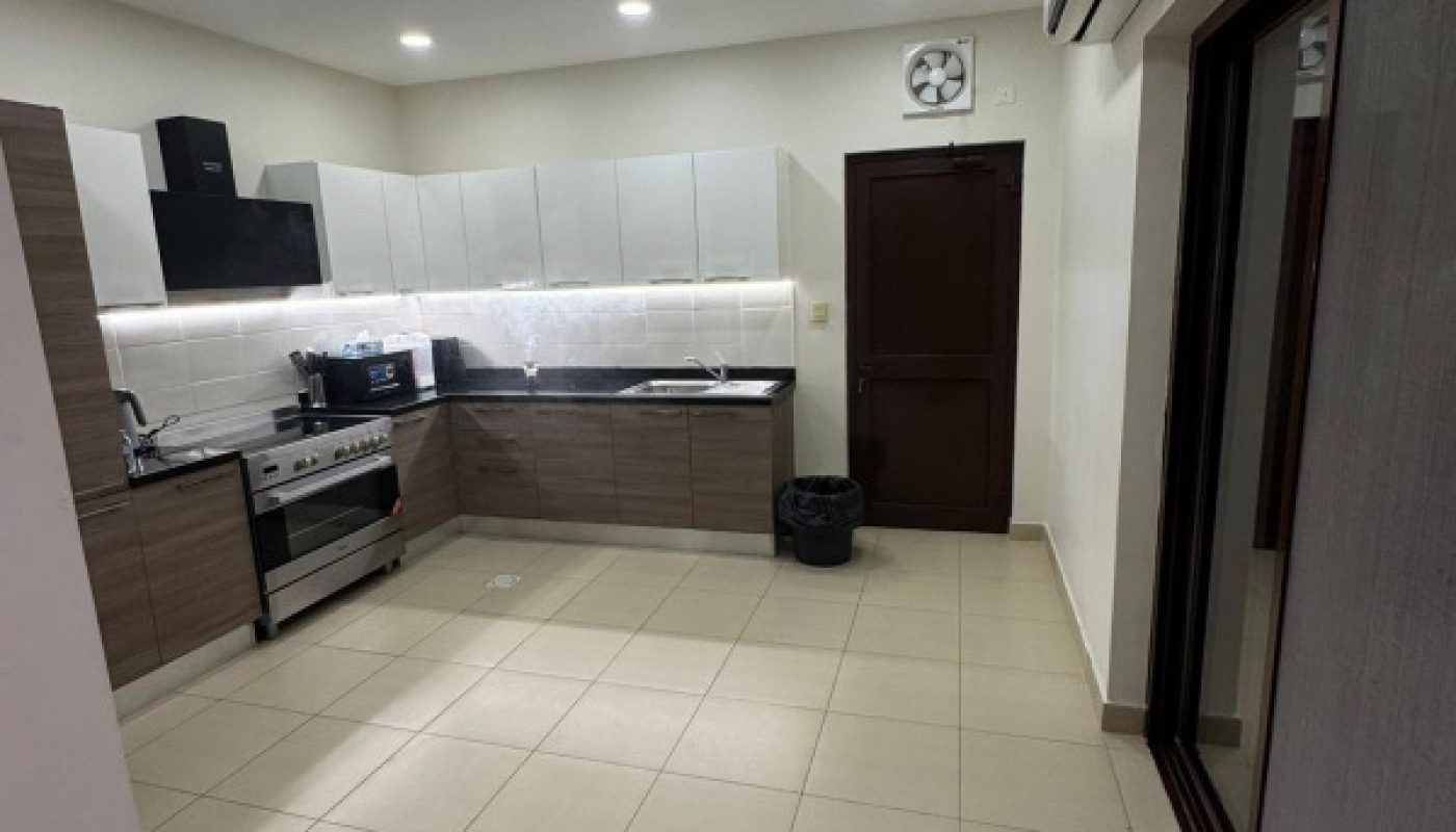 Modern kitchen with tiled floor, brown and white cabinets, black countertop, stainless steel appliances, a trash bin near a brown door, and air conditioning unit on the wall.