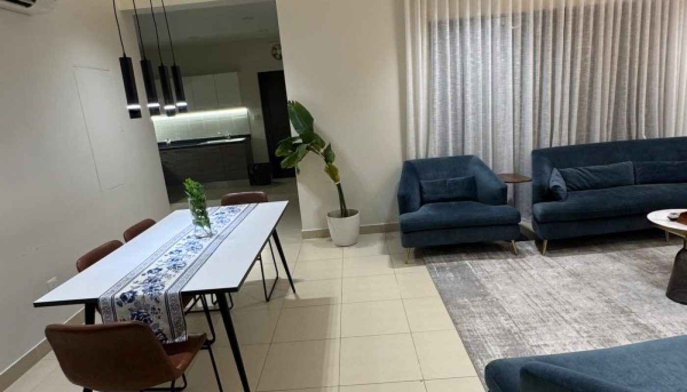 Modern living and dining area with blue sofas, brown chairs, a white table, pendant lights, a potted plant, and large curtains covering a window.
