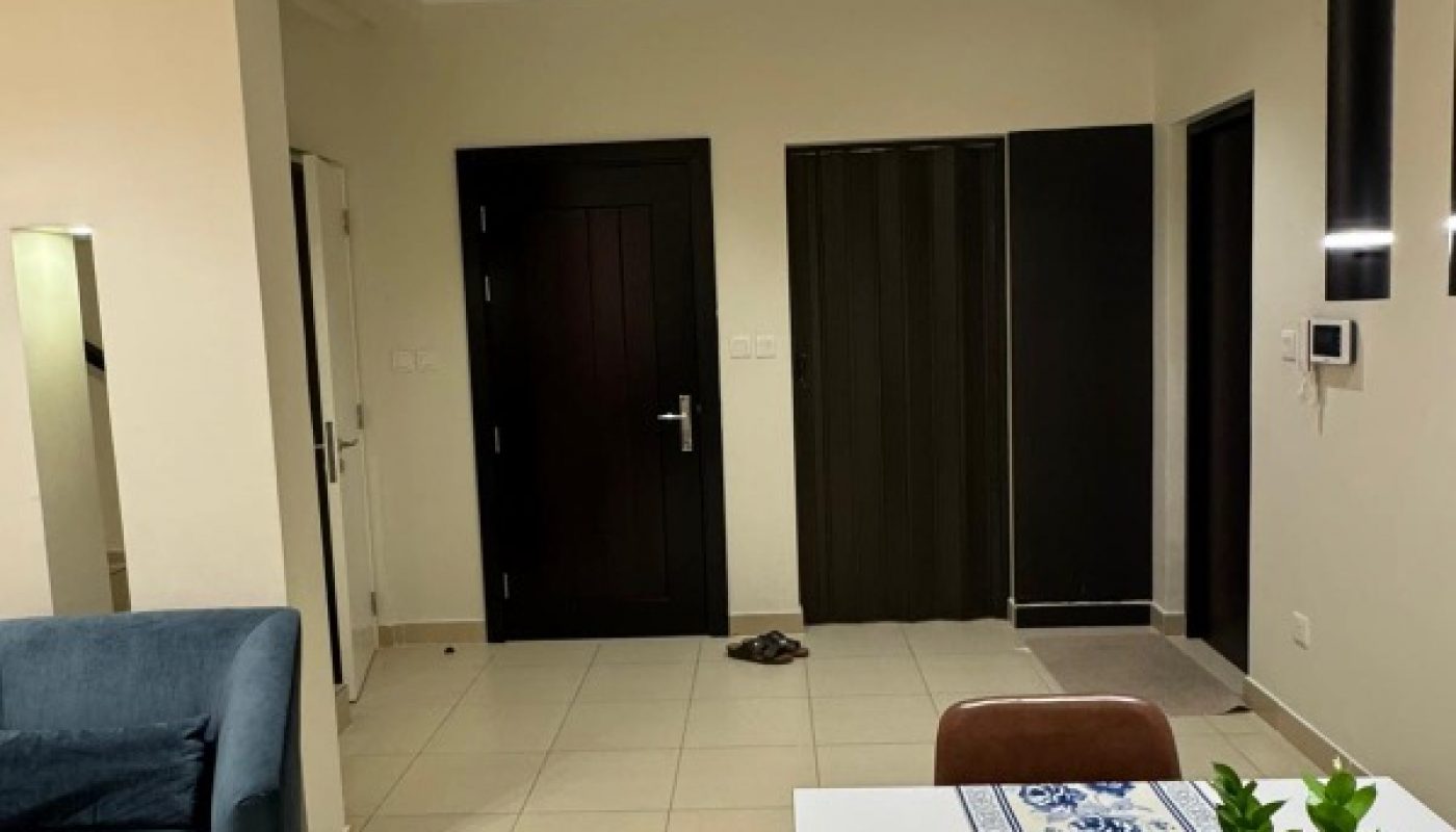 Modern living room with tiled floor, black doors, a blue sofa, a dining table with a blue-patterned runner, and a glass vase with green leaves. One shoe is on the floor near the door.