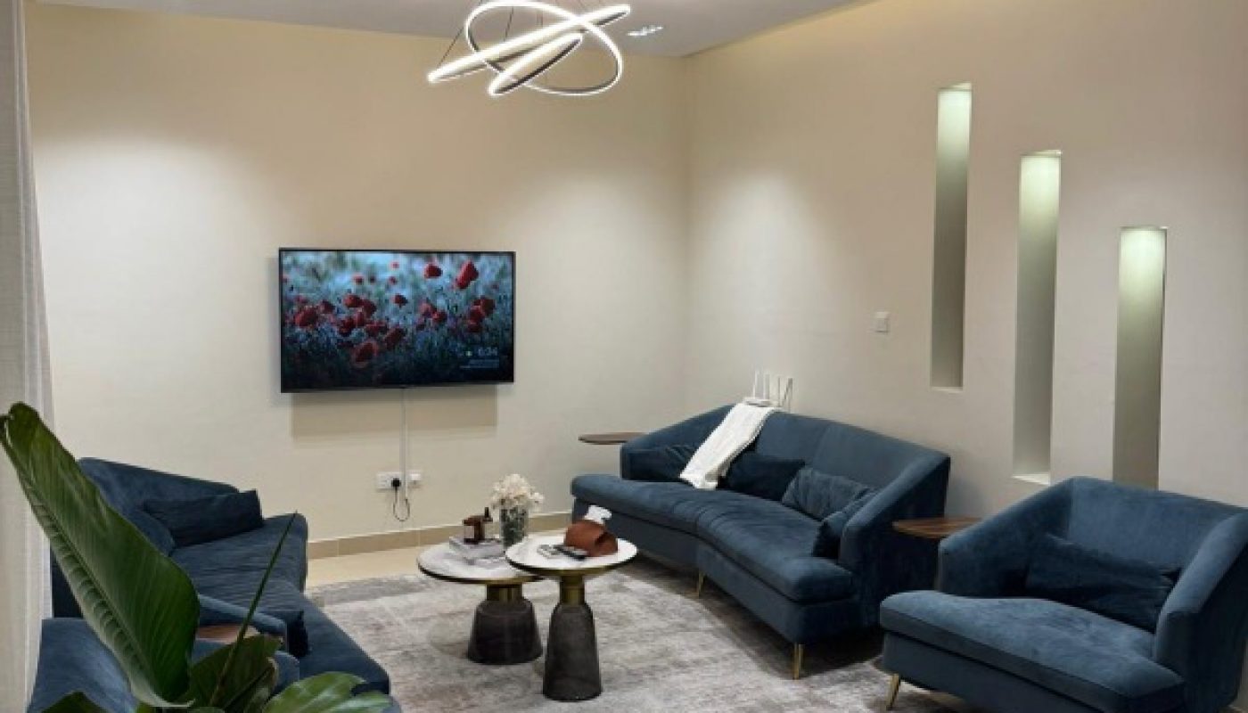 Modern living room with blue sofas, a wall-mounted TV, a round coffee table, indoor plant, abstract lighting fixture, and neutral walls with decorative vertical cutouts.