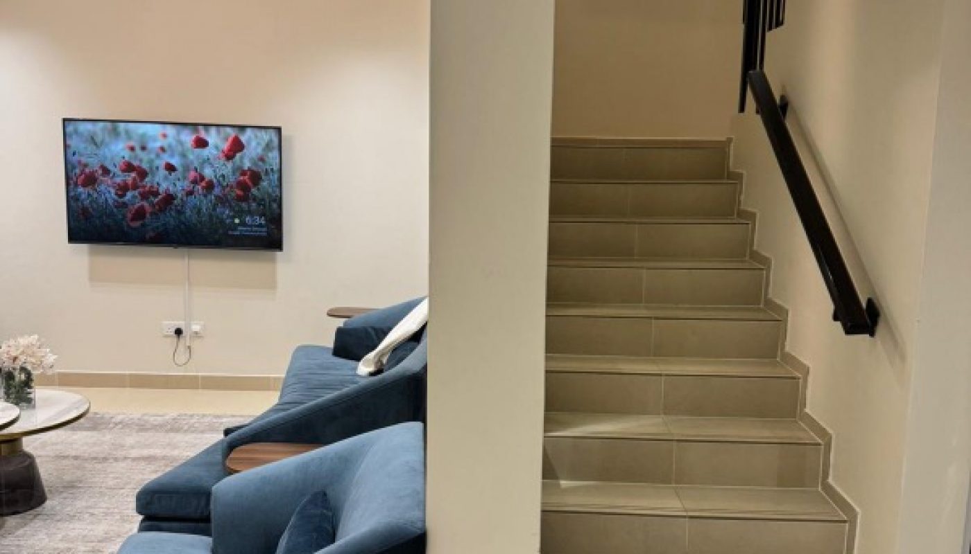 A staircase with a black handrail is beside a living area with blue sofas, a wall-mounted TV displaying flowers, and a circular chandelier.