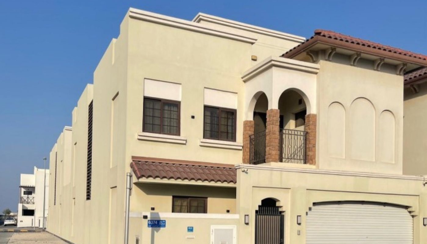 A modern beige two-story villa with a tile roof, arched balcony, closed garage door, and black gate, located on a sunny street corner.