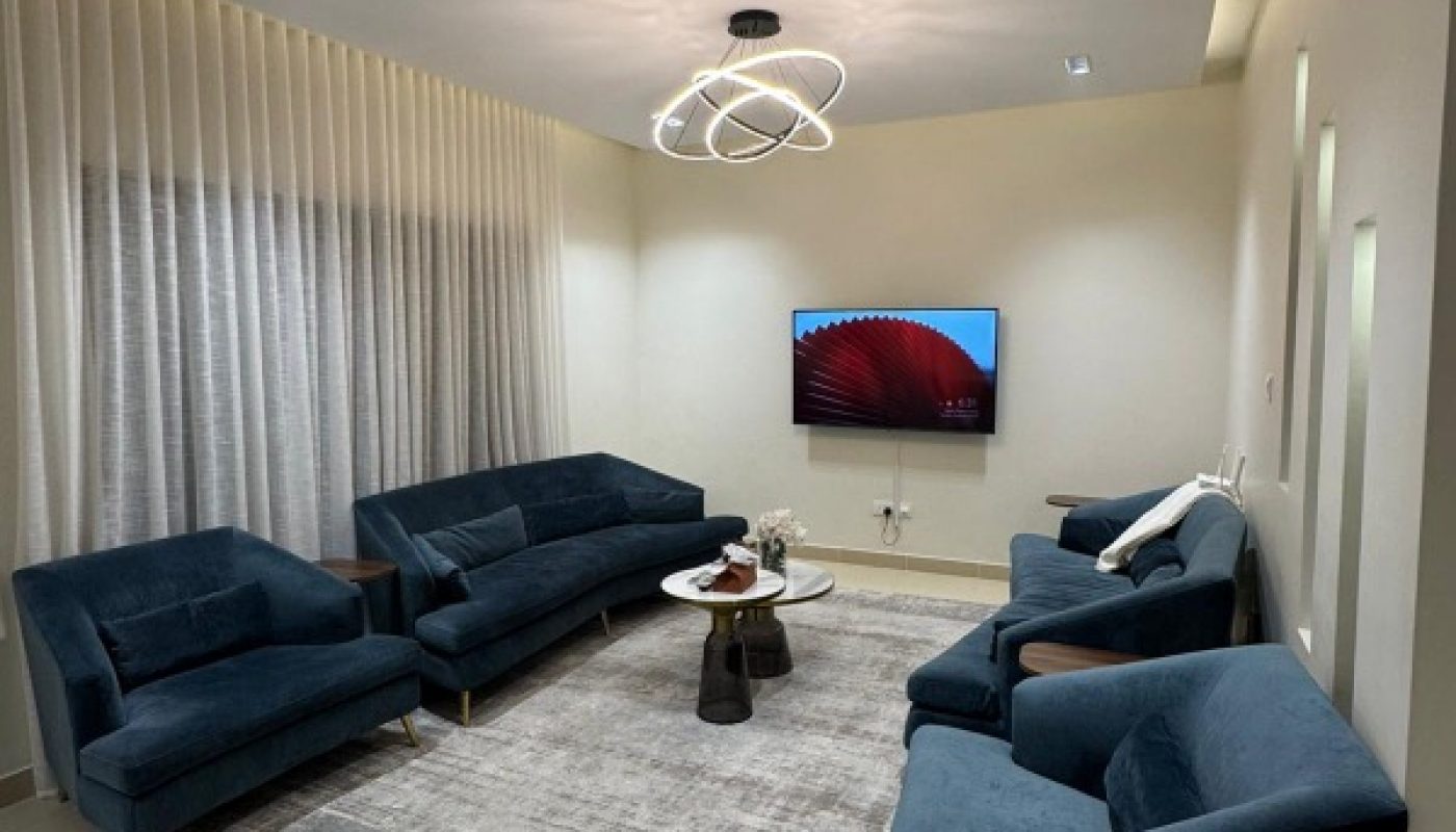 Modern living room with blue sofas, a gray rug, white walls, a TV on the wall, circular ceiling light, and floor-to-ceiling curtains on one side.