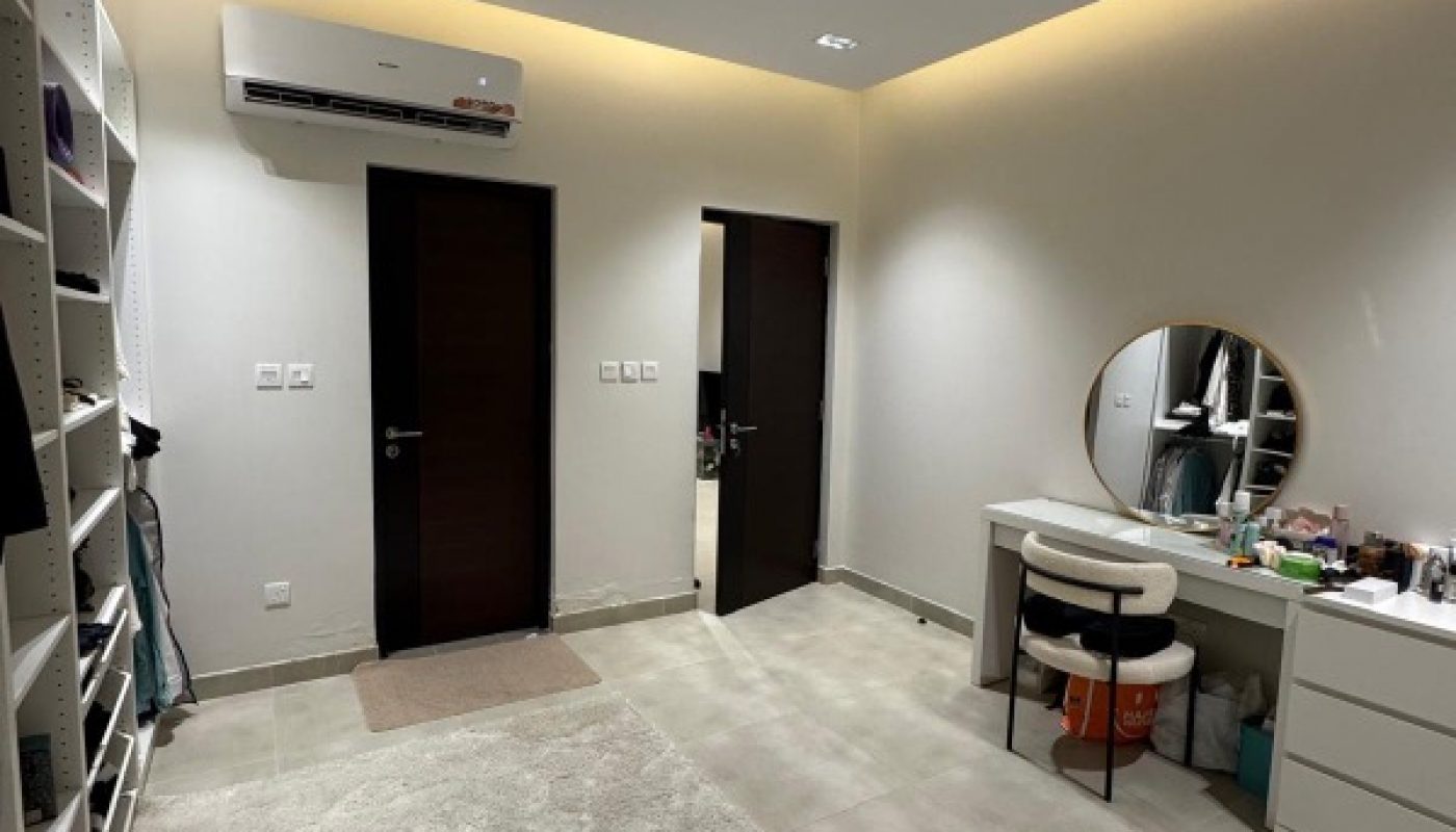 A tidy room with light-colored tile floor, two dark wooden doors, a vanity with a round mirror, a shelf, and split air conditioner above one door.