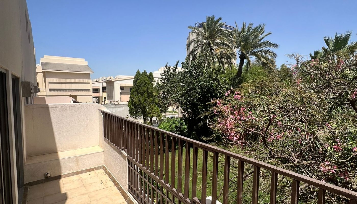 A tiled balcony with a metal railing overlooks a garden with trees and shrubs under a clear blue sky.