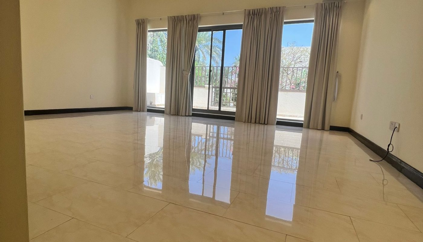 Spacious, empty living room with large cream tile floor, floor-to-ceiling windows, light curtains, and a view of trees outside.