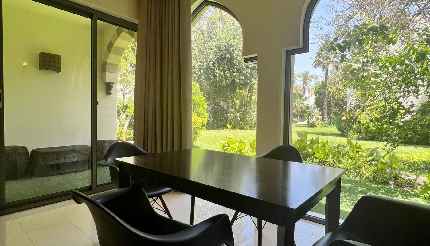 A black table with four chairs is set near large arched windows, overlooking a green garden; sliding glass doors separate another indoor space.