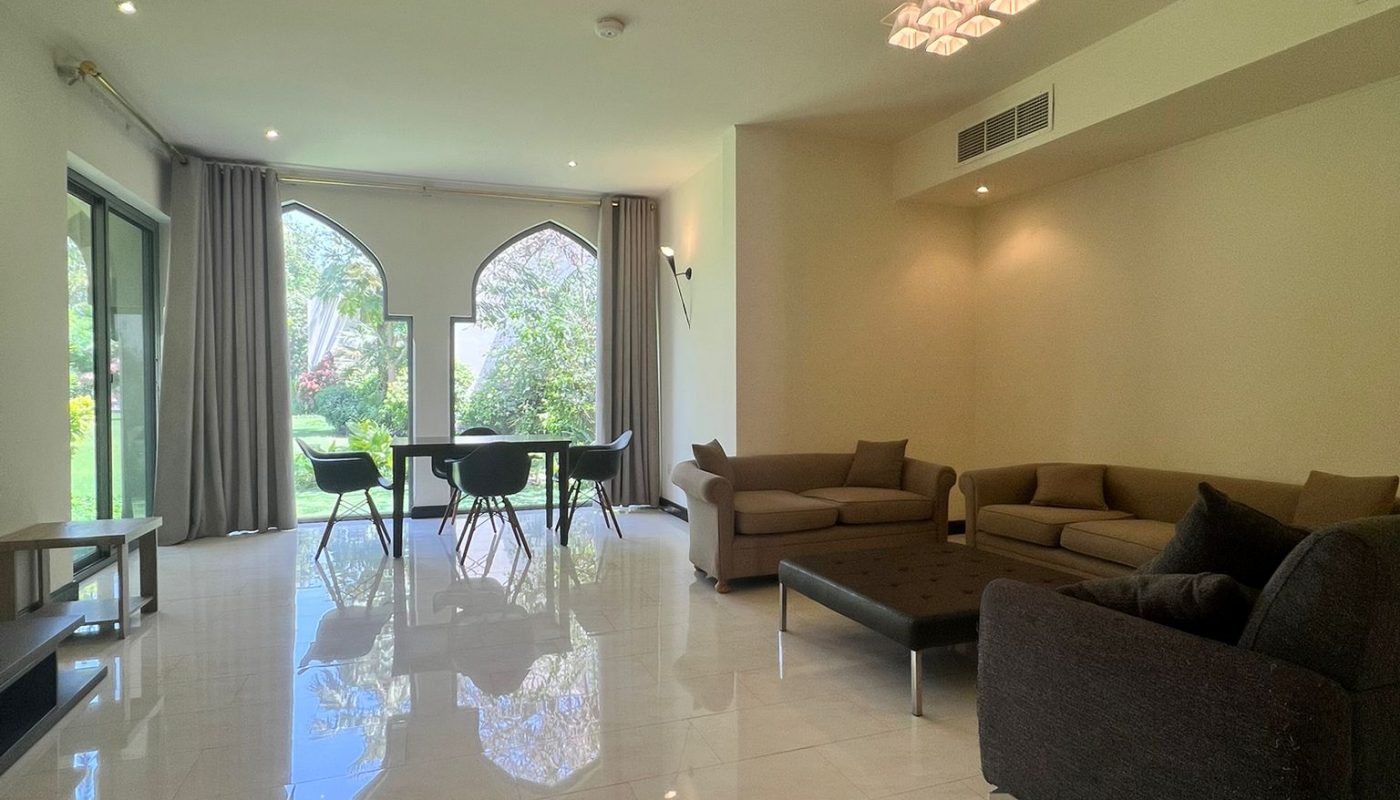 Spacious living room with tiled floor, modern sofas, a glass dining table, and large windows with garden view. Neutral-colored walls and ceiling lights are visible.