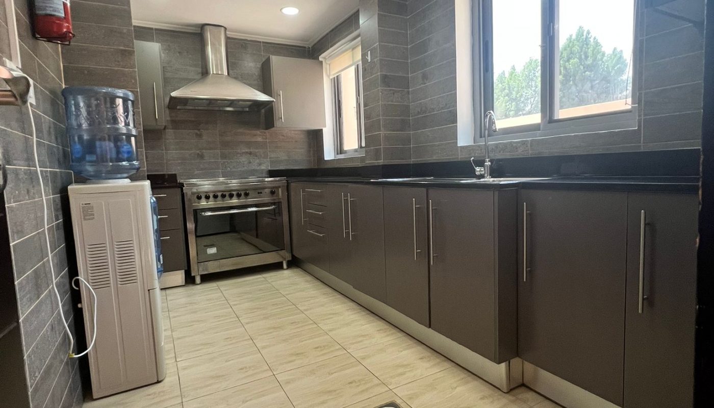 Modern kitchen with gray cabinets, stainless steel oven, water dispenser, and large windows letting in natural light. Light tiled floor and gray tiled walls are visible.
