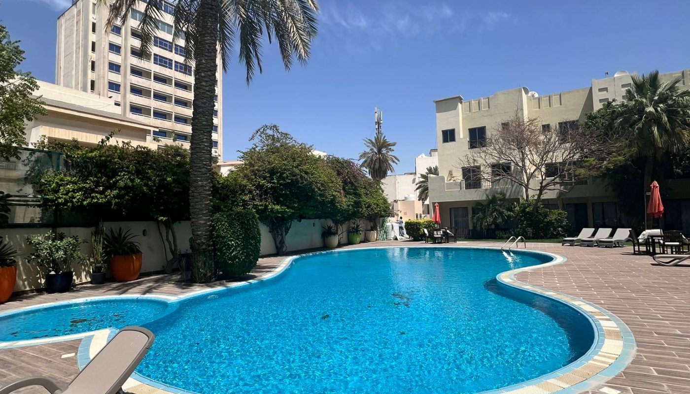 Outdoor swimming pool surrounded by lounge chairs, palm trees, and buildings under a clear blue sky.