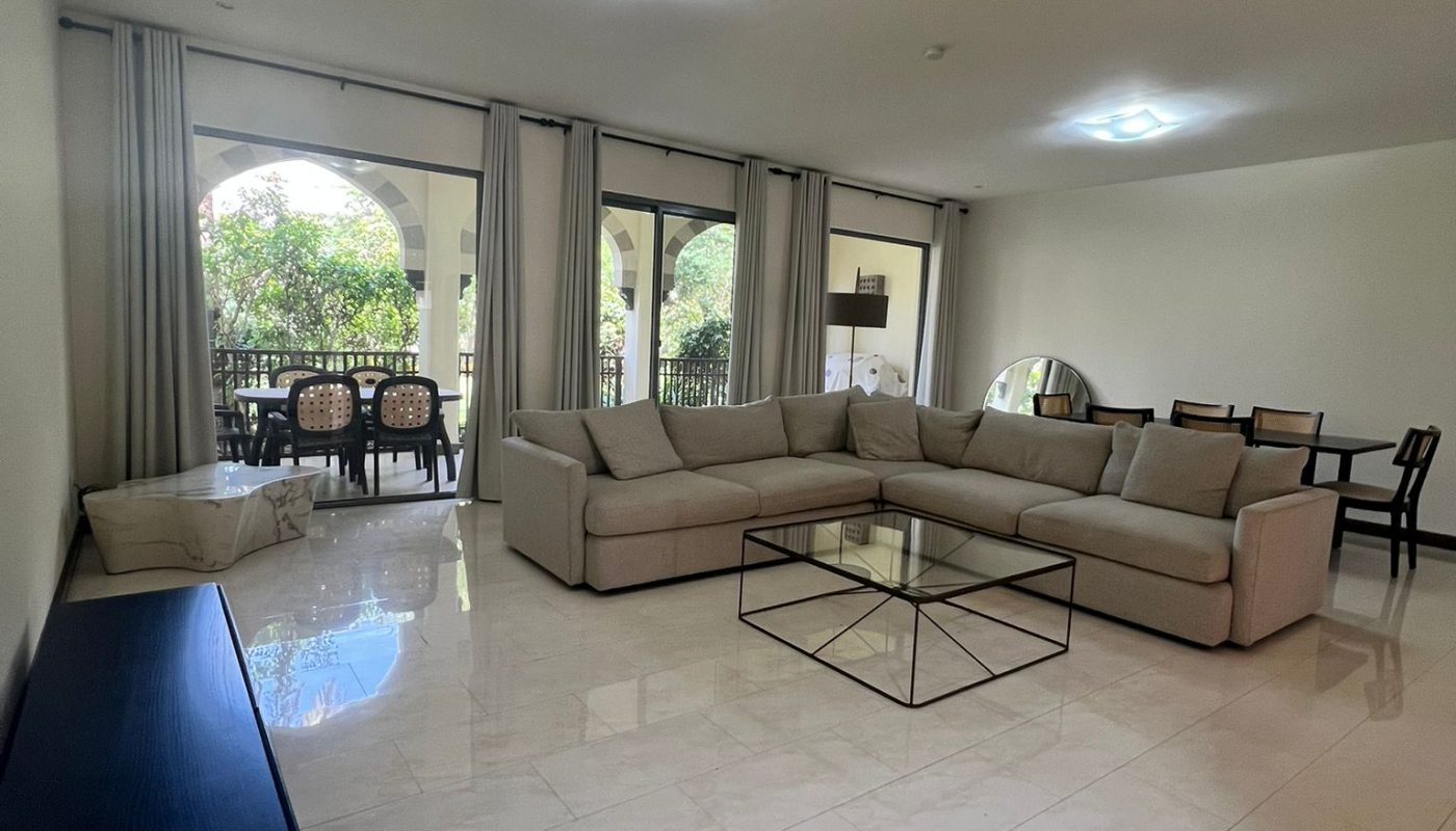Spacious living room with a large beige sectional sofa, glass coffee table, dining area, and floor-to-ceiling windows opening to a patio with outdoor seating.