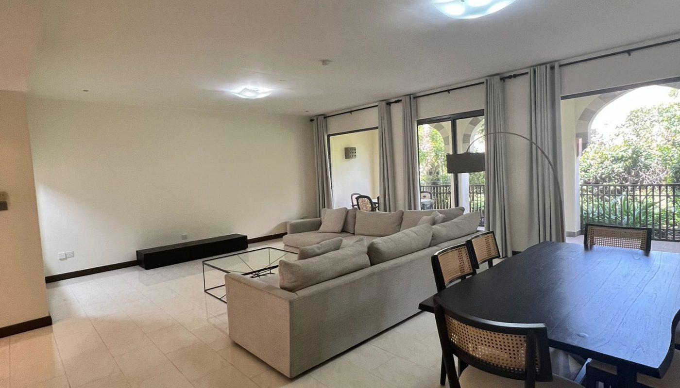 A modern living and dining area with a beige sofa, glass coffee table, dark dining table with chairs, large windows, and light tiled floors.