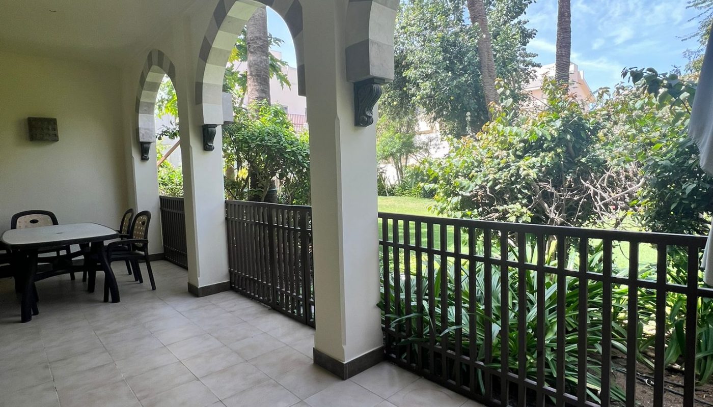 Covered patio with arched openings, tiled floor, and black railing, overlooking a garden with palm trees and greenery under a partly cloudy sky.
