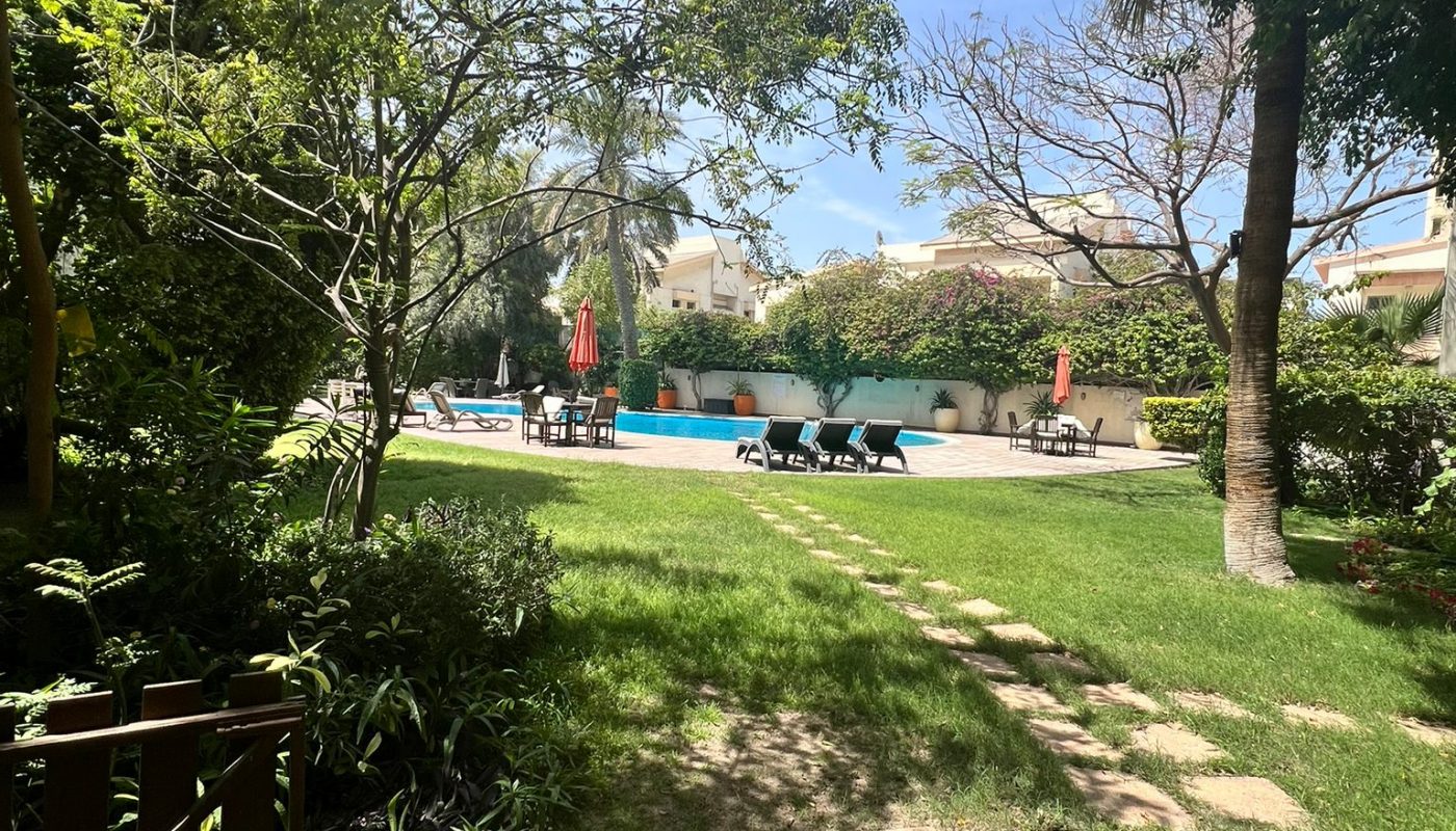 A stone path leads across a grassy yard toward a swimming pool with lounge chairs and red umbrellas, surrounded by trees and greenery under a partly cloudy sky.