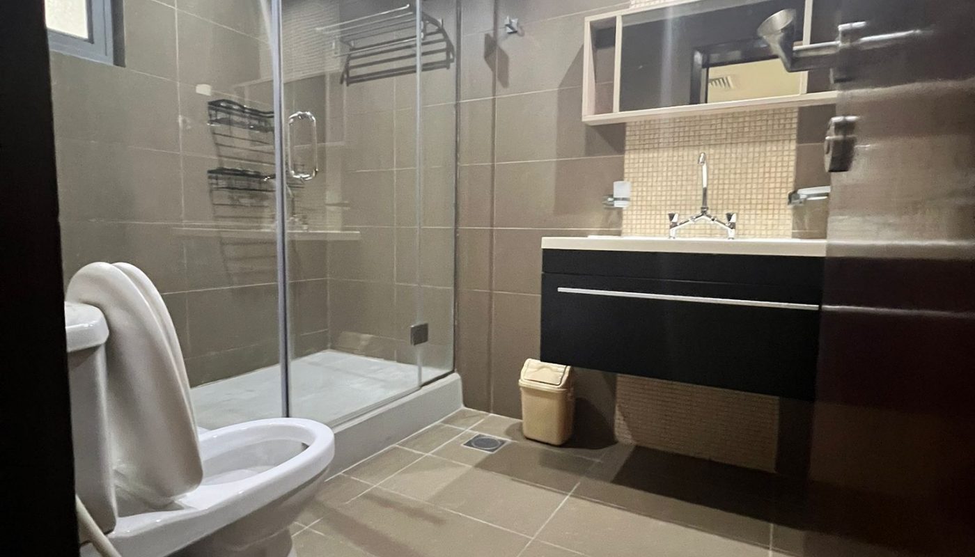 Modern bathroom with brown tiles, a glass-enclosed shower, a toilet, a vanity with sink and mirror, overhead lighting, and a small trash bin on the floor.