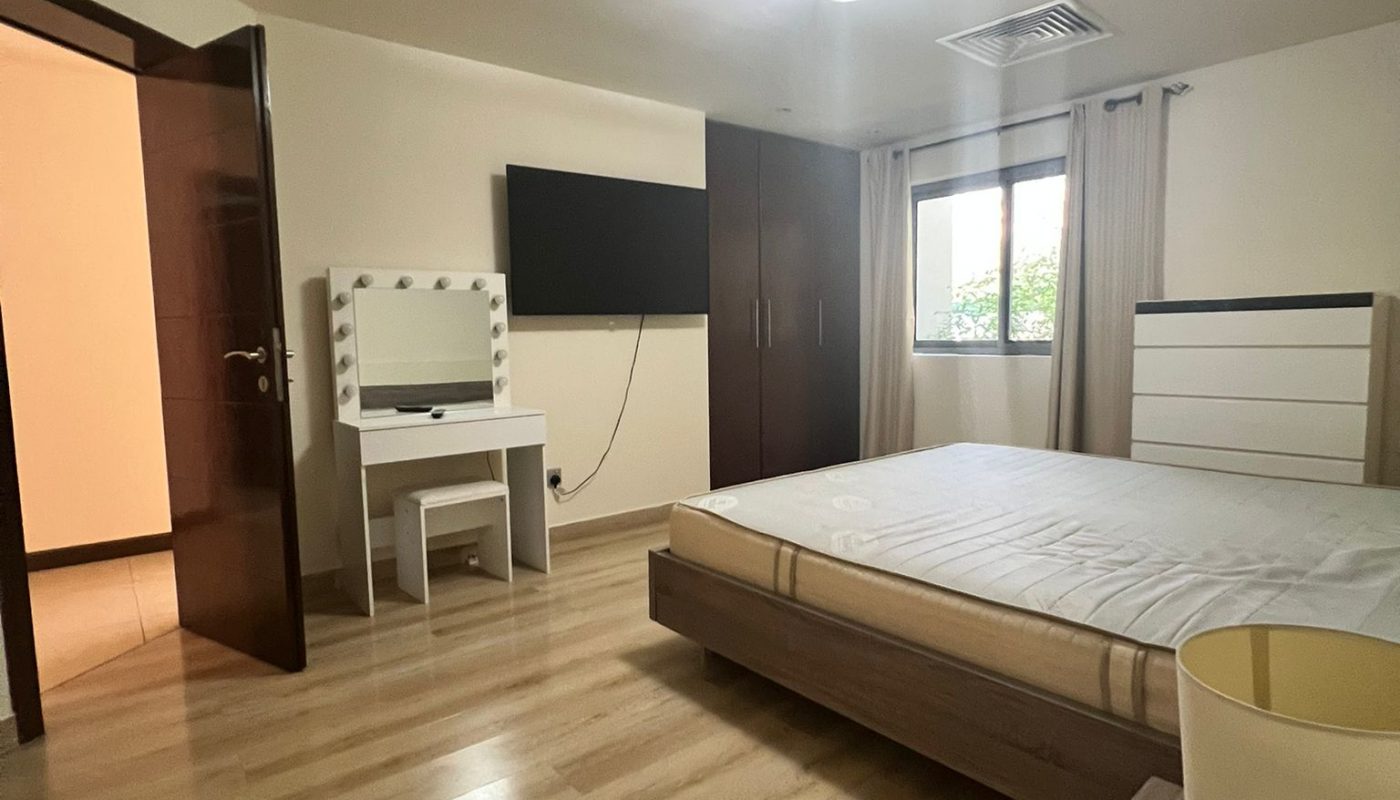 A modern bedroom with a double bed, wall-mounted TV, vanity table with mirror, dresser, and open door, featuring wood flooring and a window with curtains.