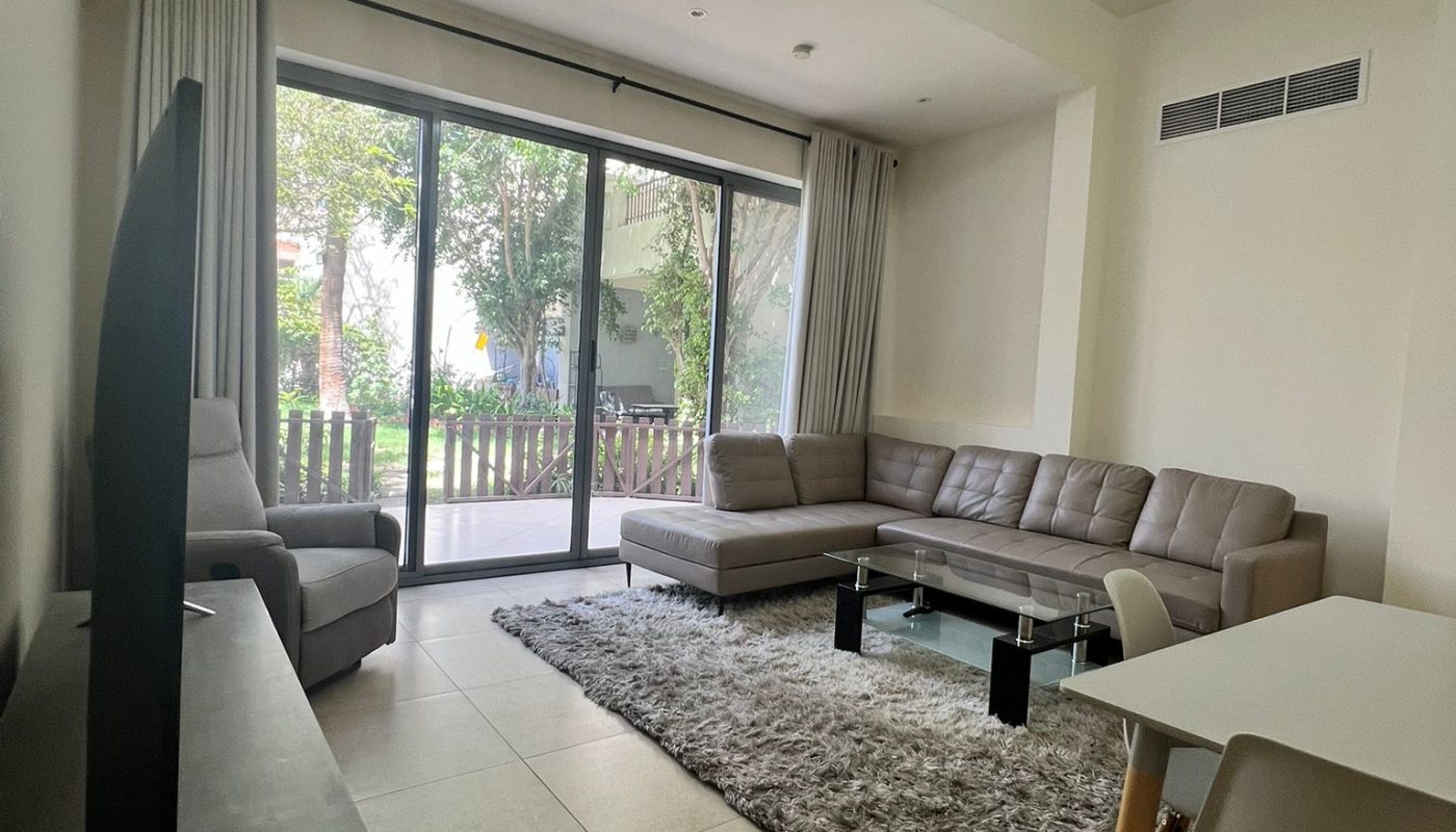 A modern living room with a gray sectional sofa, glass coffee table, TV on a stand, shaggy rug, and large sliding glass doors leading to a garden.