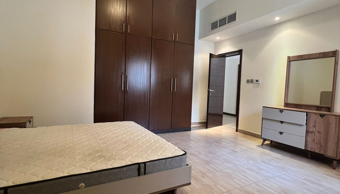 A bedroom with a bare mattress on a bed frame, a large wooden wardrobe, a dresser with a mirror, tiled floors, and neutral-colored walls.