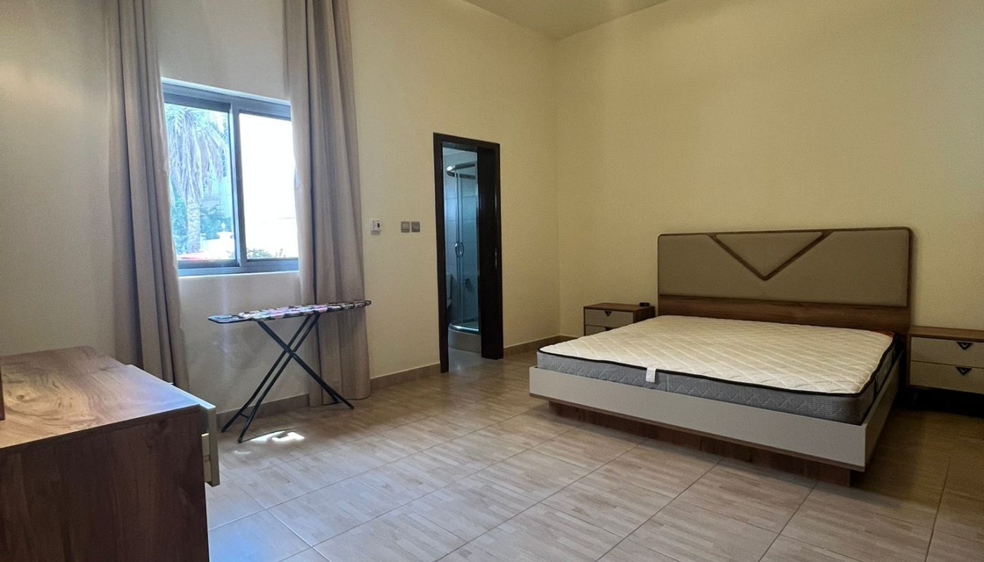 A spacious bedroom with a double bed, wooden dresser, ironing board, tiled floor, window with beige curtains, and a door leading to another room.