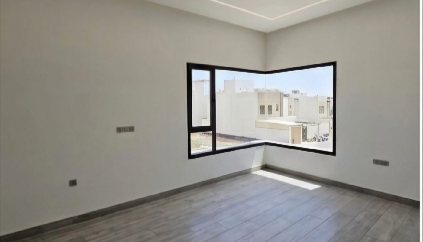 Empty modern room with gray wood-look tile flooring, off-white walls, a recessed ceiling with LED lighting, and a large corner window showing neighboring buildings.