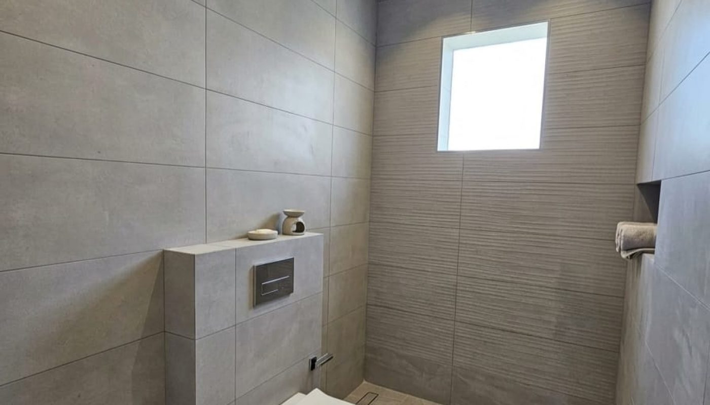 Minimalist bathroom with gray tiles, wall-mounted toilet, small window, recessed shelf with a towel, and toilet paper holder.