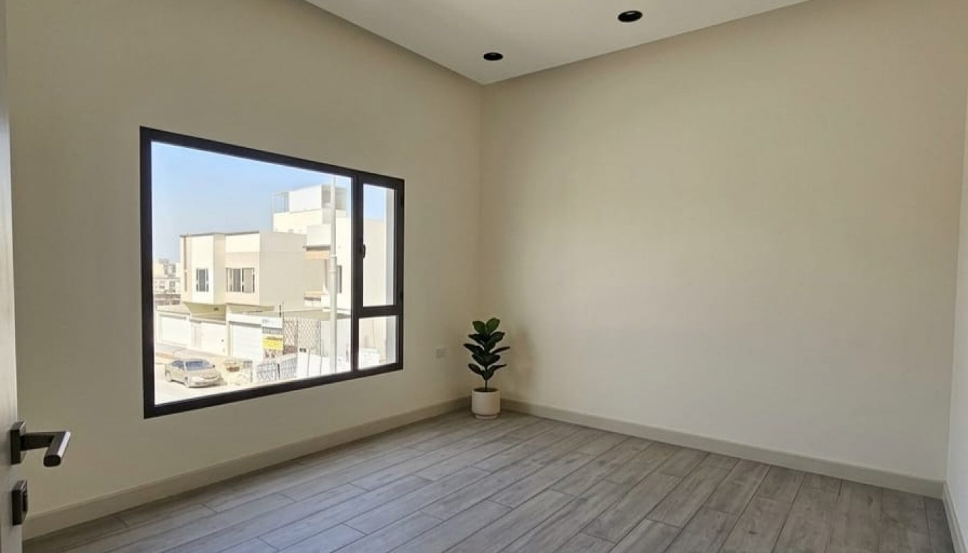 Empty, unfurnished room with light-colored walls, wood-style flooring, recessed ceiling lights, a large window, and a potted plant in the corner.