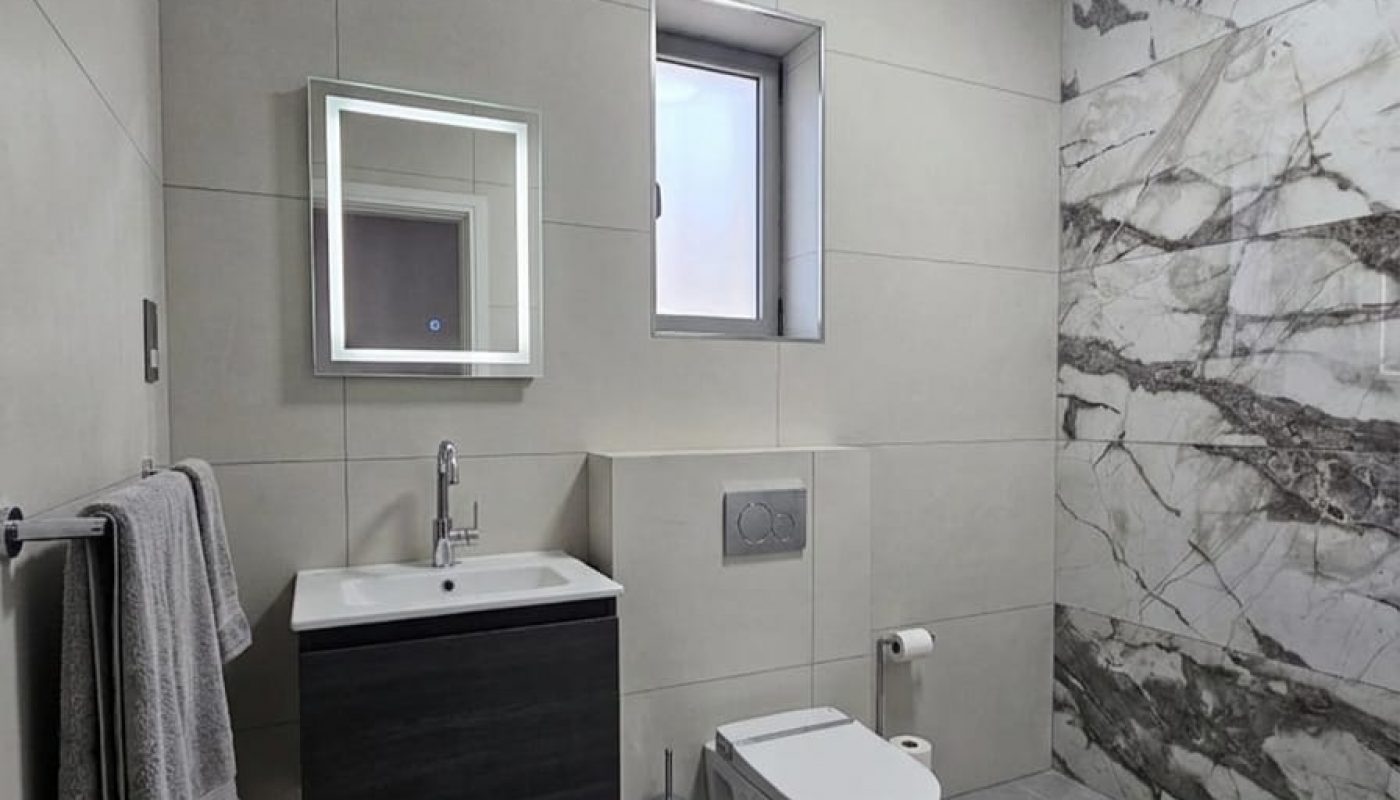 Modern bathroom with gray floor tiles, wall-mounted toilet, floating vanity with sink, illuminated mirror, towels, and a section of marble-patterned wall.