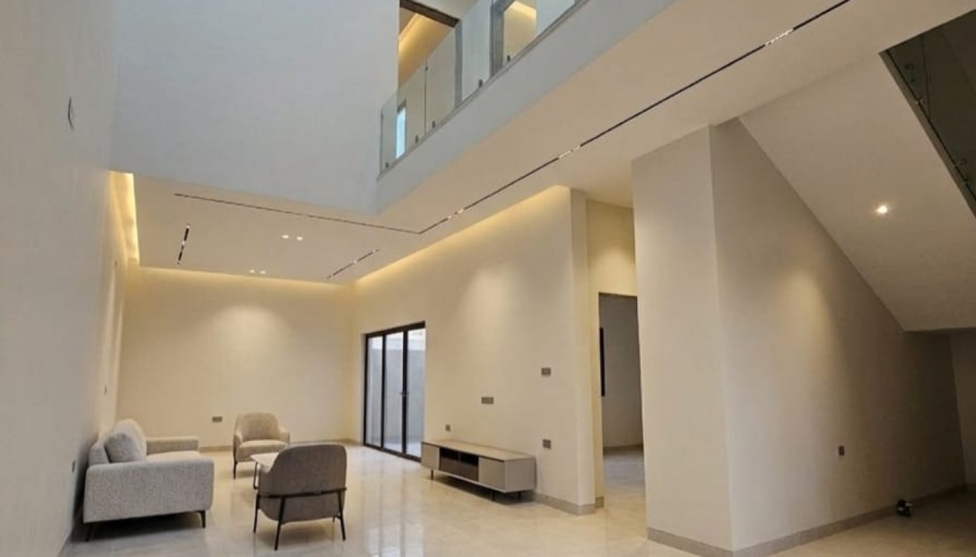 Spacious modern living area with high ceiling, skylight, neutral-toned walls and floors, minimal furniture, and glass balcony on the upper level.