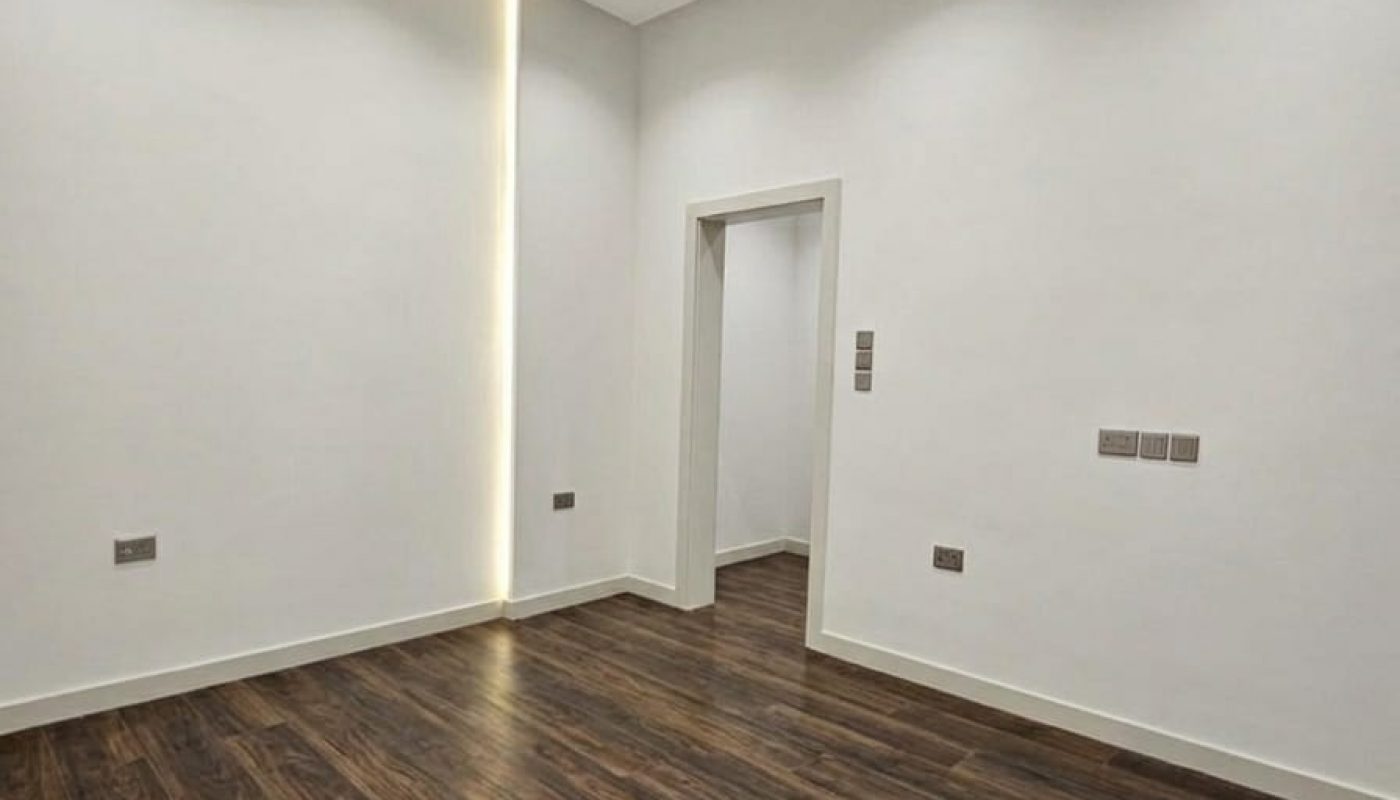 Empty room with white walls, recessed ceiling lights, dark wood flooring, a narrow vertical light strip on one wall, and a doorway leading to another space.