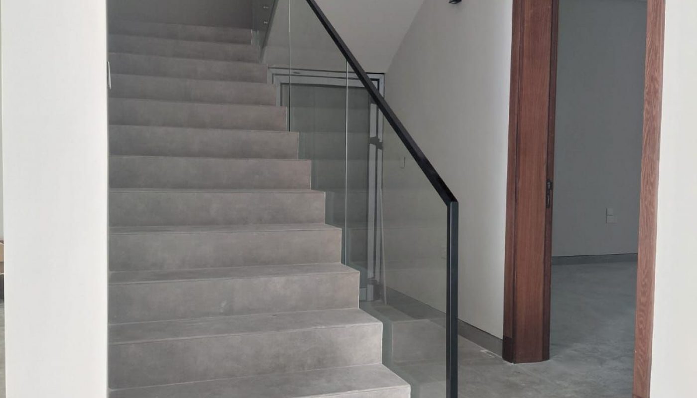 Concrete staircase with glass and metal railing ascending to an upper floor, next to a doorway with wooden trim, in a modern interior space with gray tile flooring.