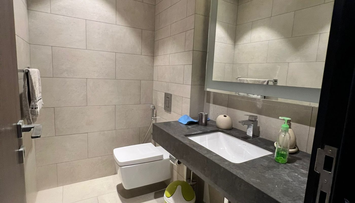 Modern bathroom with beige tiled walls, a floating toilet, a countertop sink with a large mirror, a green trash bin, and various toiletries on the counter.