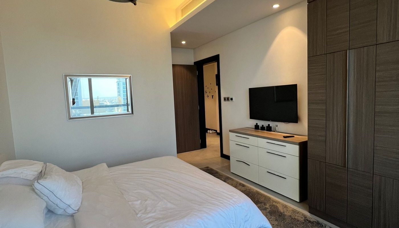A modern bedroom with a neatly made bed, a wall-mounted TV on a cabinet, a large wardrobe, and a ceiling light fixture, featuring neutral decor and soft lighting.