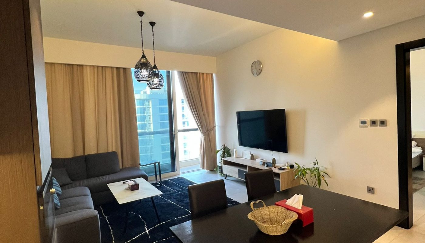 Modern living and dining area with a black dining table, dark sofa, wall-mounted TV, large rug, and floor-to-ceiling windows with beige curtains.