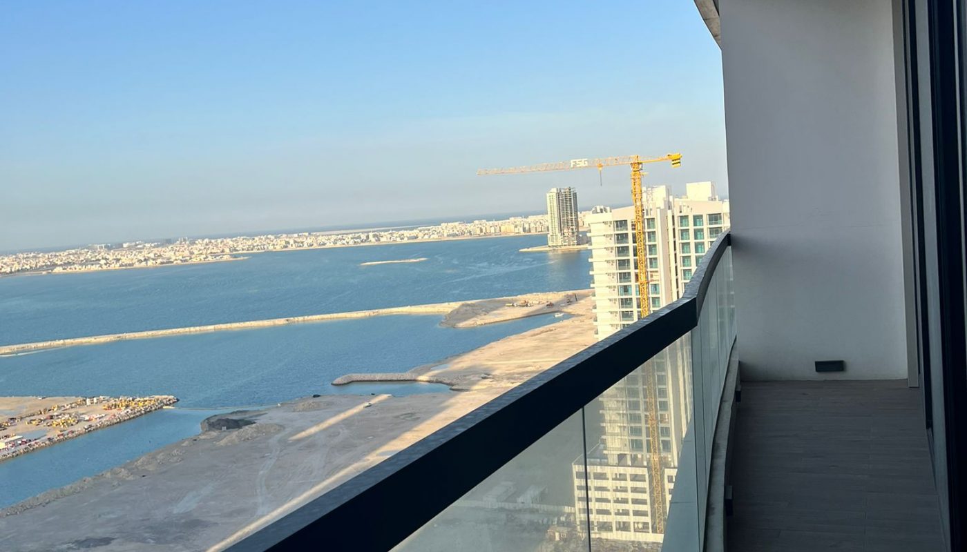View from a high balcony overlooking a waterfront, construction cranes, modern buildings, and clear blue sky in the background.