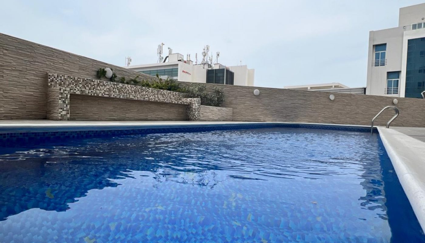 Rectangular outdoor swimming pool with blue mosaic tiles, surrounded by stone walls and buildings in the background under a cloudy sky.