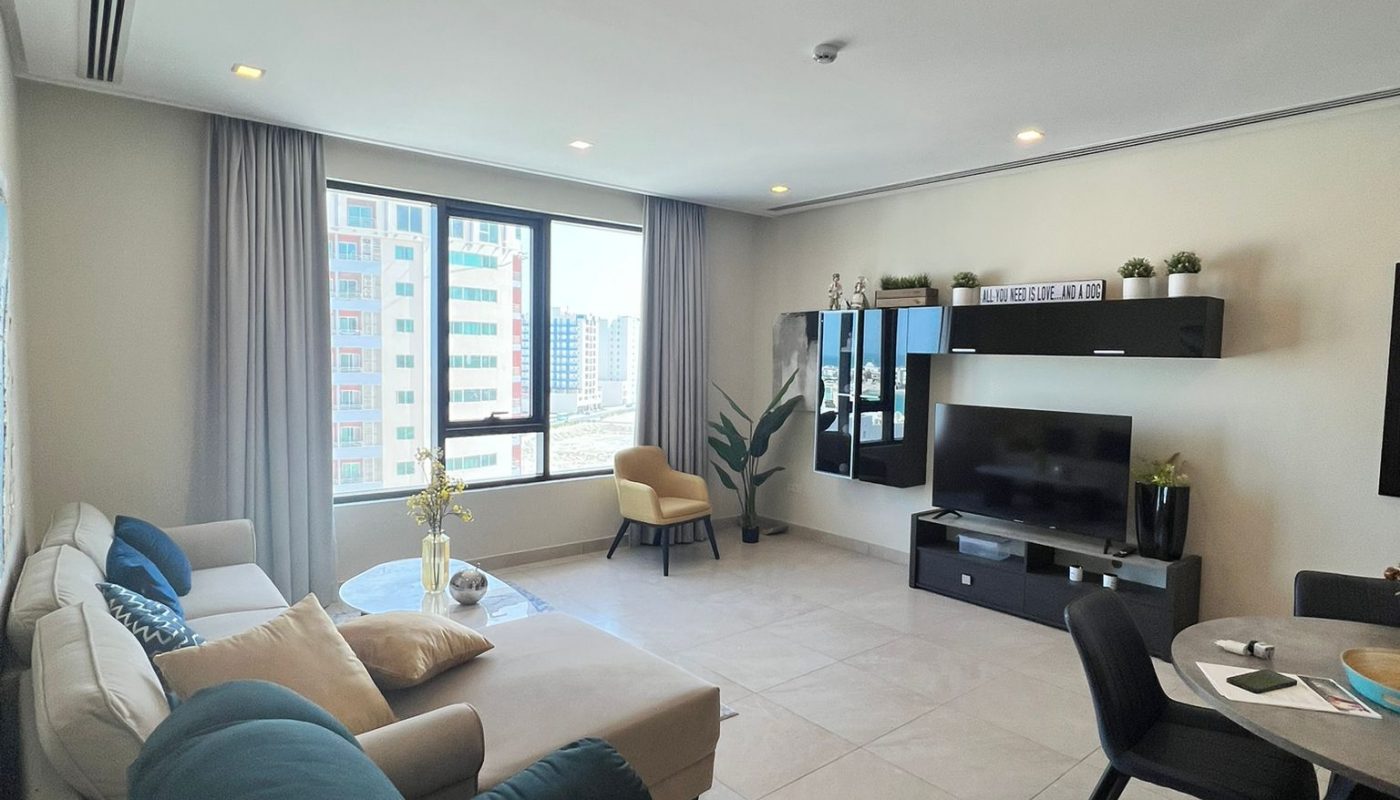 Modern living room with large windows, a sectional sofa, armchair, TV, shelving unit, dining table, and city view outside. Neutral colors and natural light fill the space.