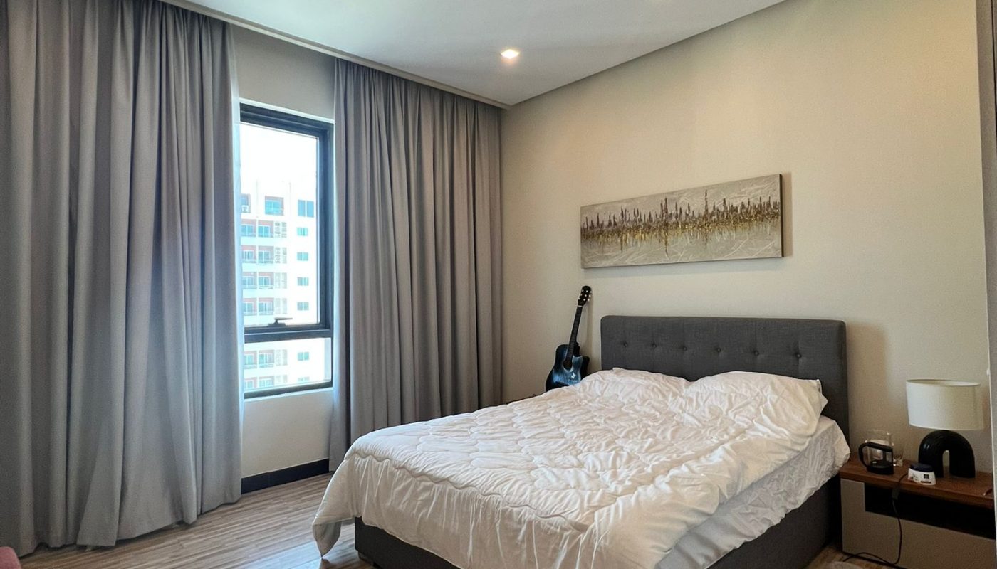 A modern bedroom with a large bed, messy white bedding, wooden floor, guitar in the corner, window with closed curtains, and a cityscape painting on the wall.