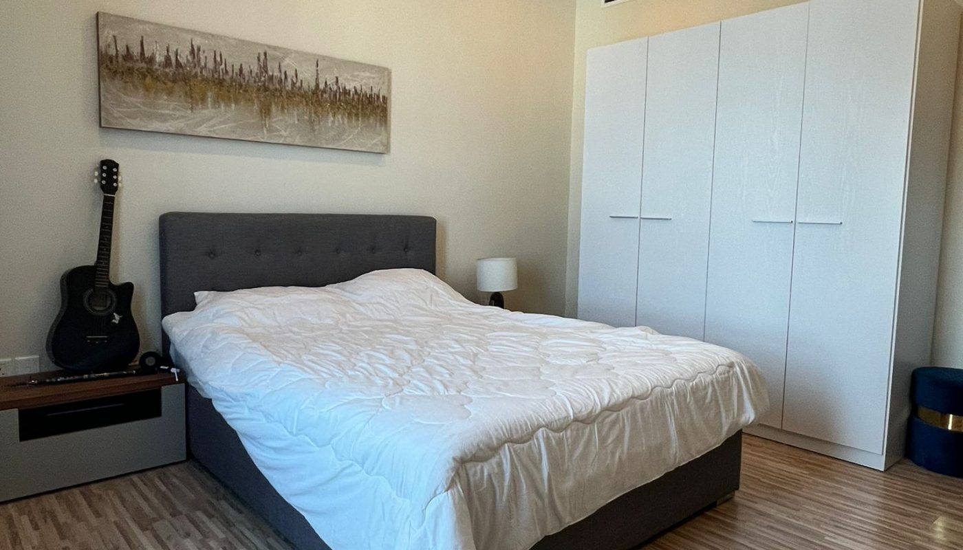 A neatly made bed with a white comforter in a modern bedroom, featuring a wardrobe, bedside table with a lamp, a guitar, and wall art above the bed.