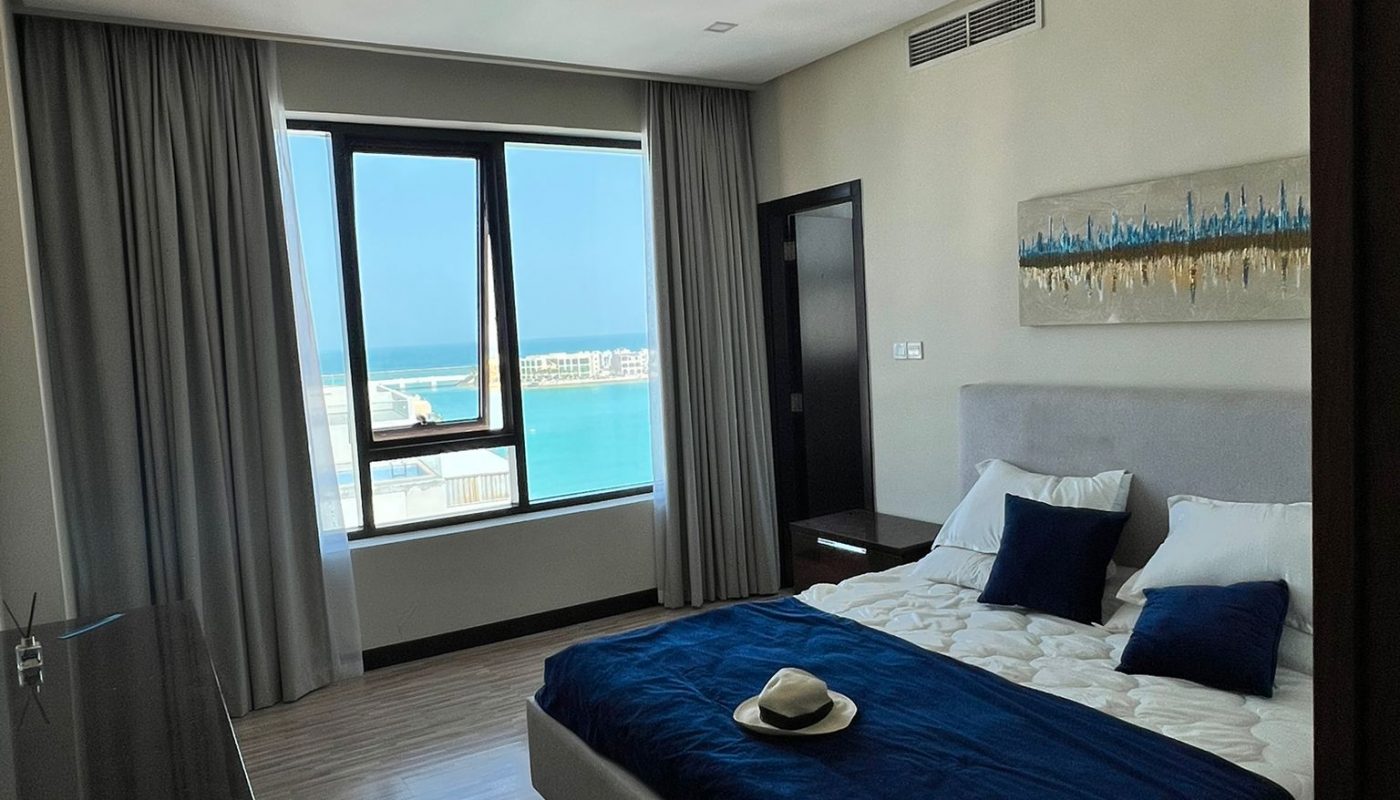A modern bedroom with a bed covered in a blue blanket, pillows, and a hat; large window reveals a view of the sea and a marina outside.