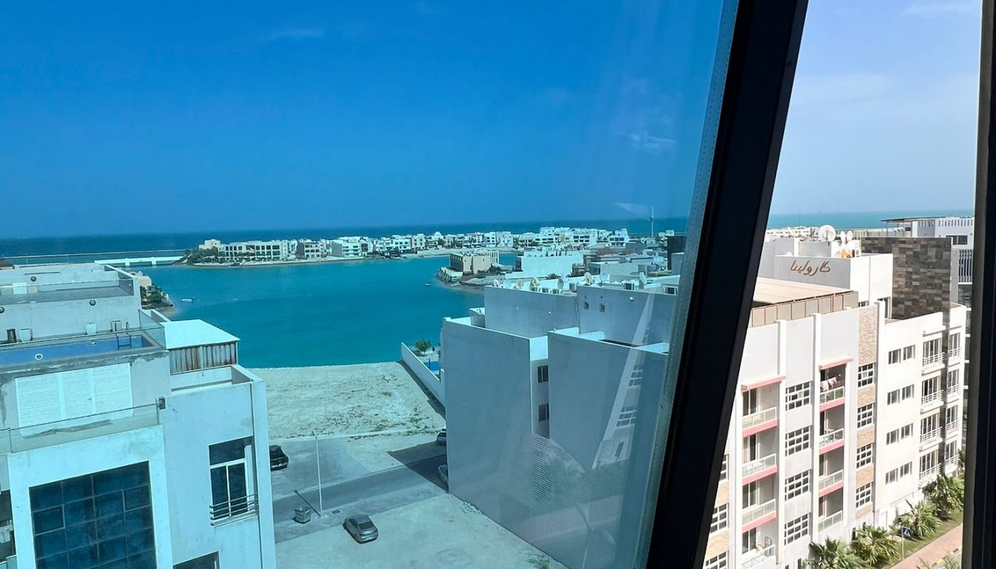 A view through an open window shows modern white buildings, a road lined with palm trees, and a blue body of water under a clear sky.