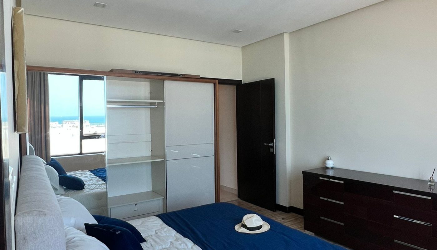 A modern bedroom with a made bed, blue accents, a hat on the bed, a dresser, mirrored closet doors, and a window showing an outdoor view.