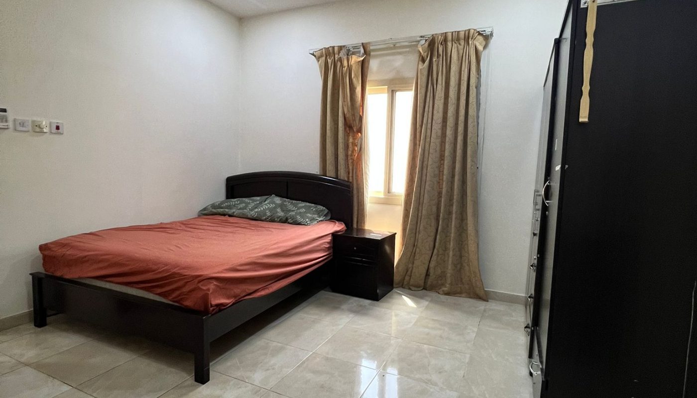 A simple bedroom with a double bed, red bedding, a nightstand, wardrobe, ceiling fan, curtains, tiled floor, and a window letting in natural light.