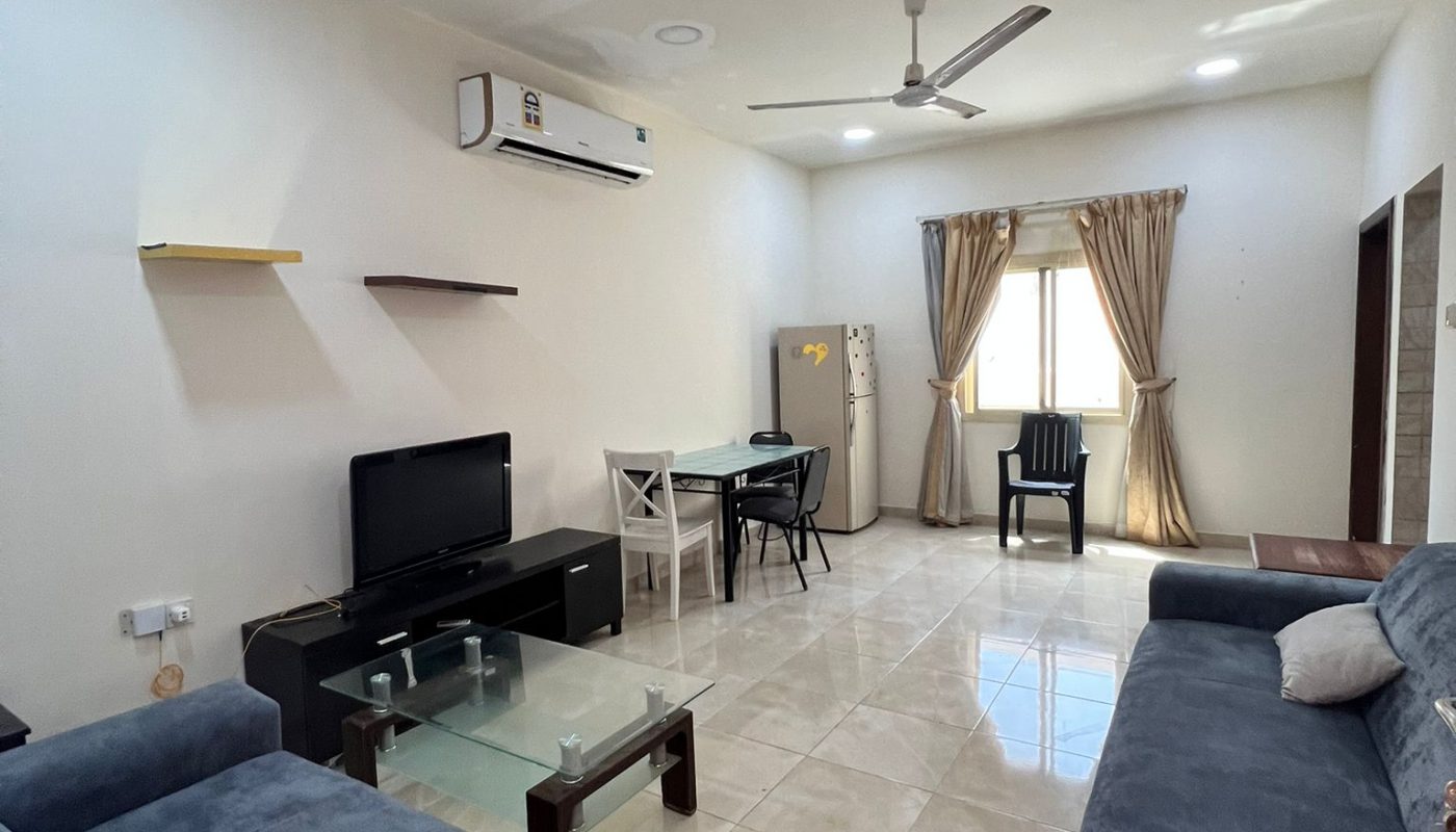A living room with two blue sofas, a TV on a stand, glass coffee table, dining table with two chairs, fridge, and a window with beige curtains.