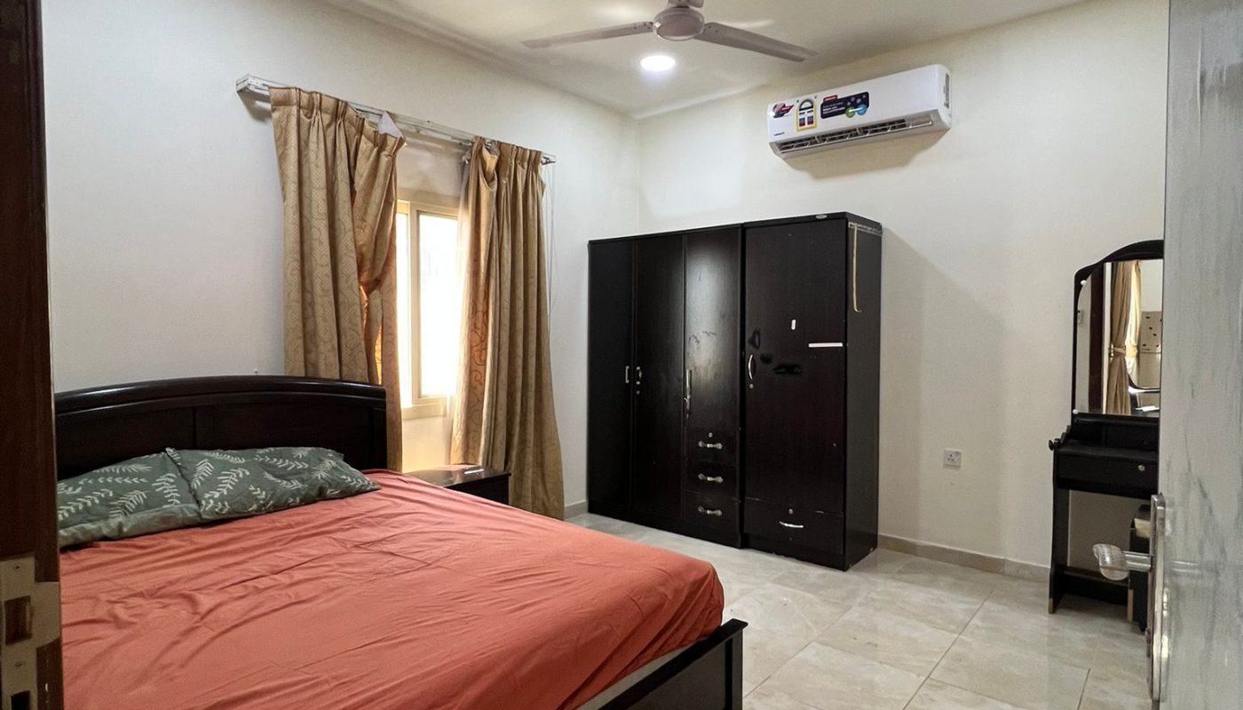 A bedroom with a double bed, orange bedding, dark wood furniture, a wardrobe, dressing table, ceiling fan, air conditioner, and tiled floor. Light comes in through a curtained window.