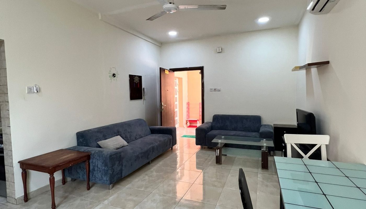 A living room with blue sofas, a wooden side table, glass dining table with black and white chairs, ceiling fan, and tiled floor. An open door leads to another brightly lit room.