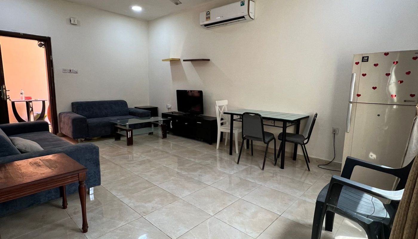 Spacious living room with tiled floor, sofas, a TV, dining table with chairs, refrigerator, ceiling fan, and air conditioner. Natural light enters from a door on the left.