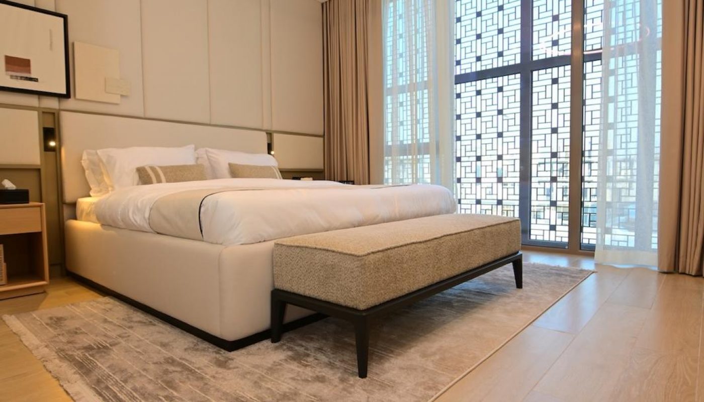 Modern bedroom with a large bed, upholstered bench, area rug, wooden floor, circular ceiling light, and floor-to-ceiling windows with patterned screens and sheer curtains.