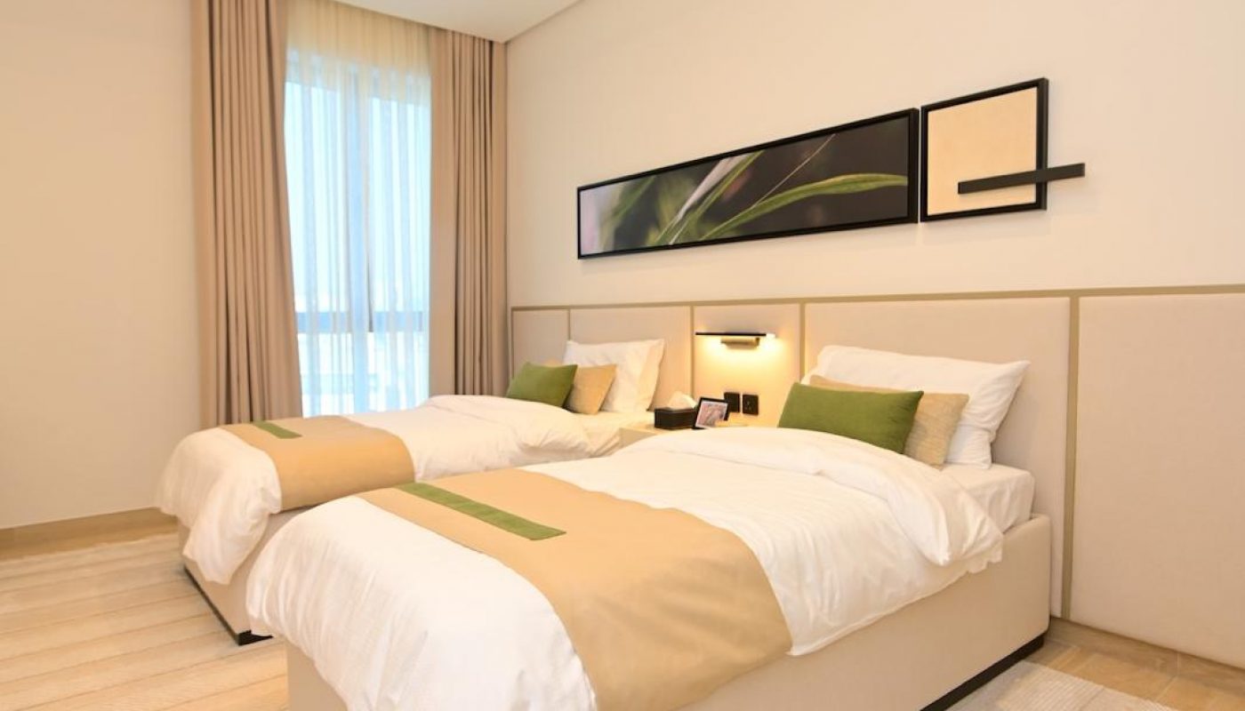 Modern bedroom with two single beds, beige bedding, green accent pillows, a large circular ceiling light, artwork above the headboards, and a window with floor-length curtains.